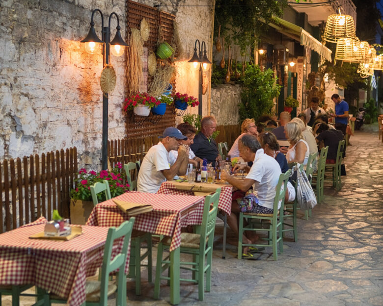 Pythagoreio is a small town and former municipality on the island of Samos, North Aegean, Greece. Greek cuisine is a Mediterranean cuisine. Contemporary Greek cookery makes wide use of vegetables, olive oil, grains, fish, wine, and meat. People in the Image are tourists enjoying the food.