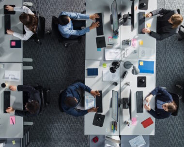 Top Down View of Diverse Colleagues Working In Corporate Office of Hedge Fund. Using Desktop Computers, Discussing Documents, Analyzing Data. Concept of Teamwork and Professional Business Environment