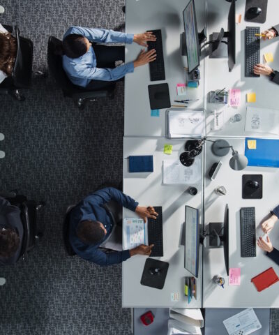 Top Down View of Diverse Colleagues Working In Corporate Office of Hedge Fund. Using Desktop Computers, Discussing Documents, Analyzing Data. Concept of Teamwork and Professional Business Environment