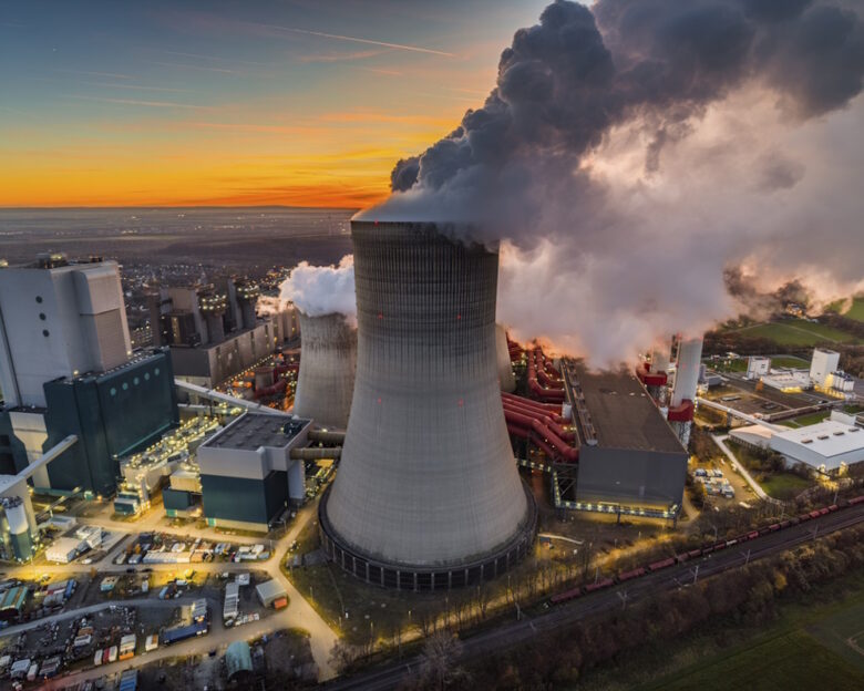 Aerial view of a large cooling tower releasing steam at an industrial power plant during sunset. Dramatic evening light and rising vapor highlight large-scale electricity generation, heavy industrial infrastructure, and energy production facilities.