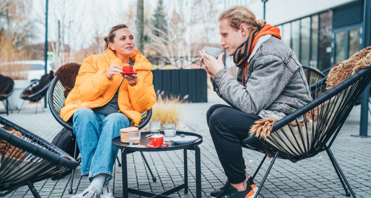 Mother with adult son sitting on the open terrace in a coffee shop, drinking coffee and having a lovely conversation. Late autumnal days in Europe.