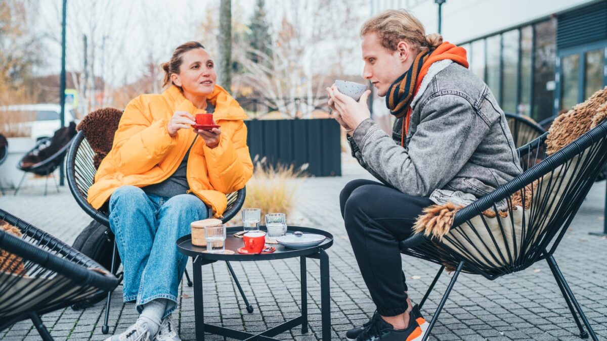 Mother with adult son sitting on the open terrace in a coffee shop, drinking coffee and having a lovely conversation. Late autumnal days in Europe.