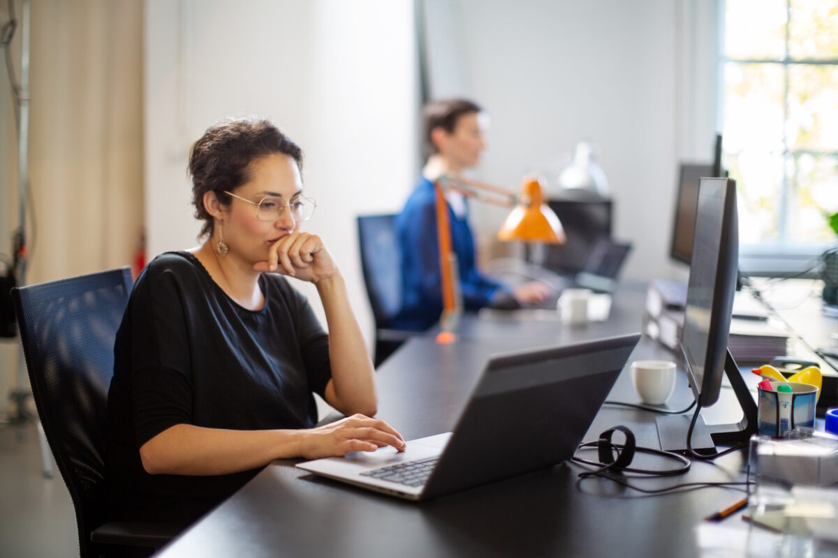 Businesswoman working on laptop with colleague in background. Female executive sitting at her desk looking busy while working on laptop computer.