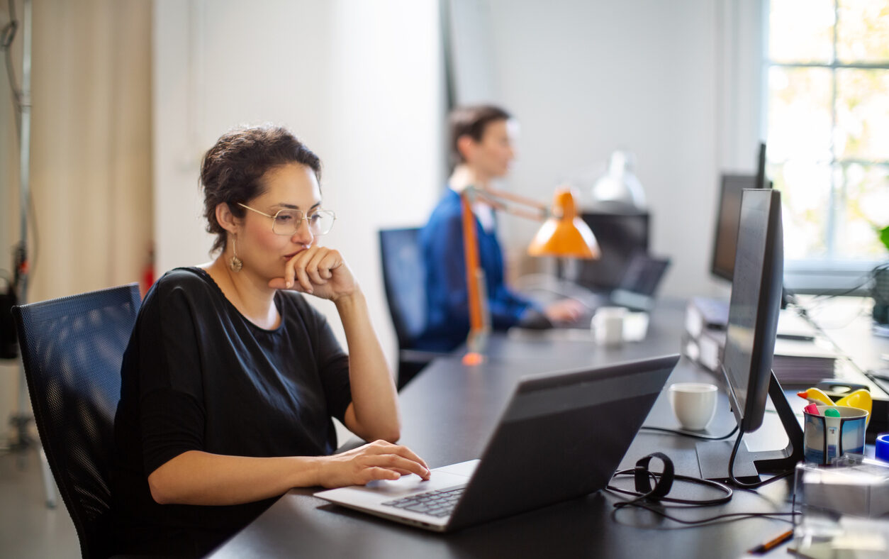 Businesswoman working on laptop with colleague in background. Female executive sitting at her desk looking busy while working on laptop computer.