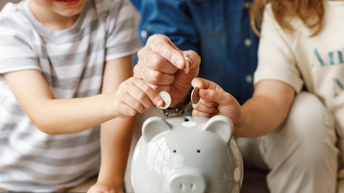 Unrecognizable senior man and kids putting coins into piggy bank on table at home together