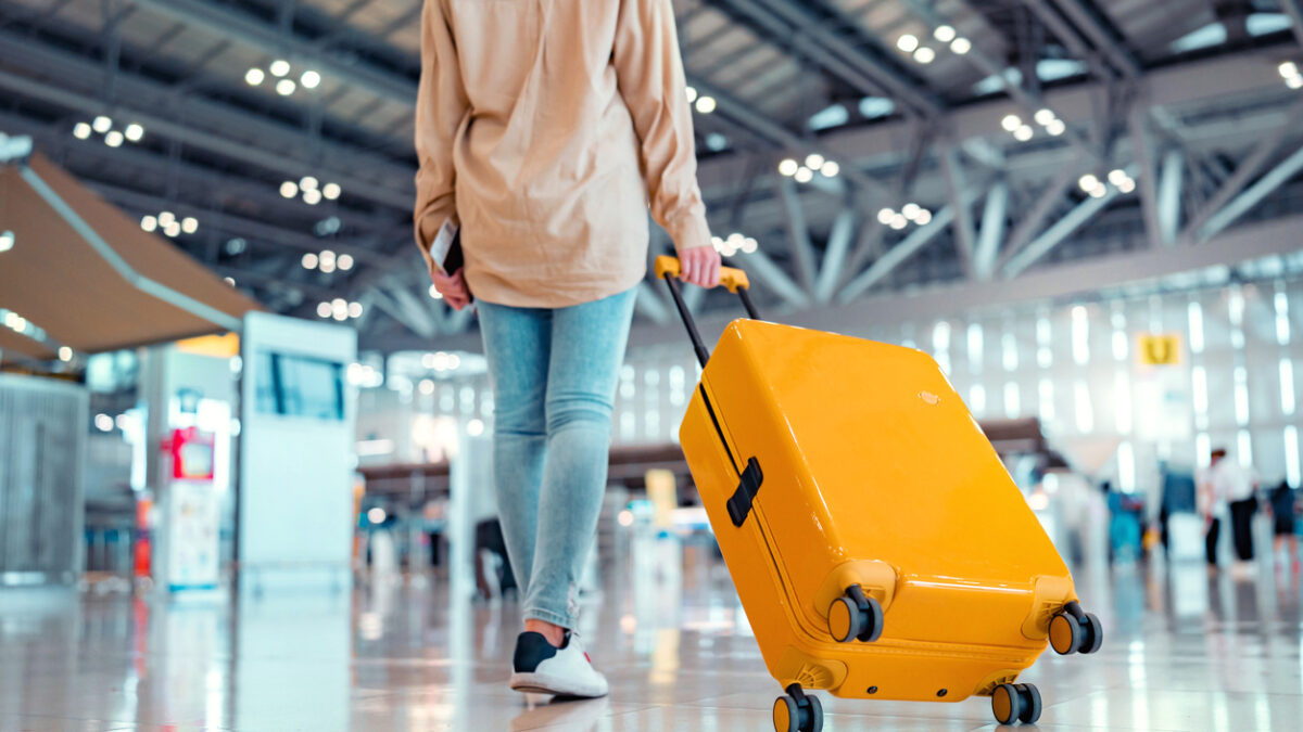 Young female traveler passenger walking with a yellow suitcase at the modern Airport Terminal, Back view of woman on her way to flight boarding gate, Ready for travel or vacation journey