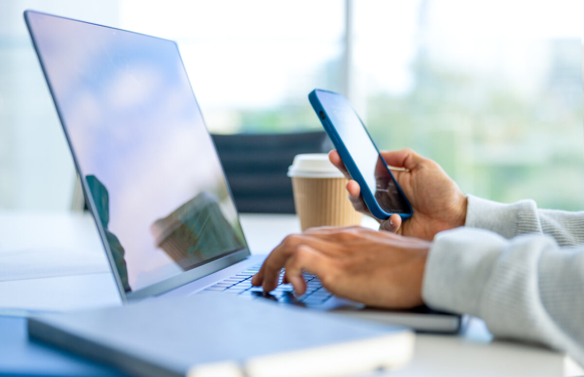 Close up of a Businessman working on a laptop computer and holding and looking at a mobile phone in the office. There are notepads on the desk. He is casually dressed