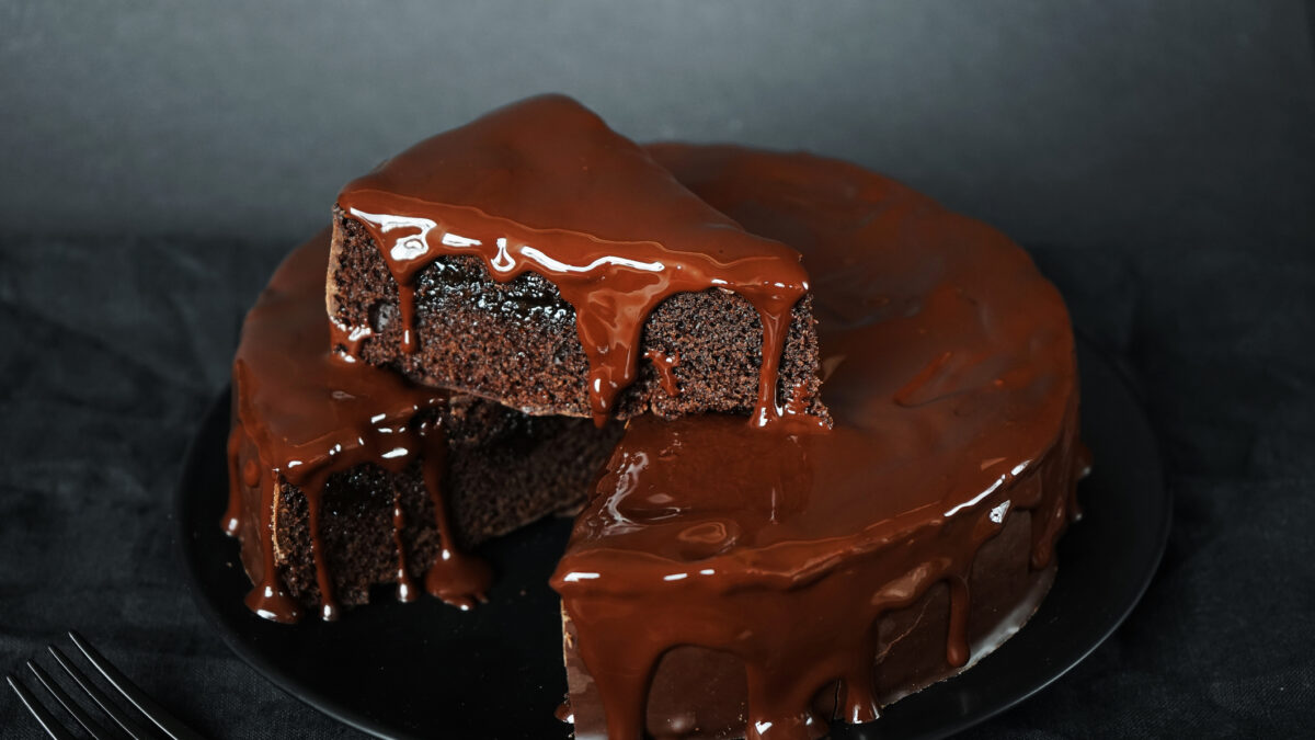 Chocolate cake with chocolate icing on a black plate next to a fork on a dark background