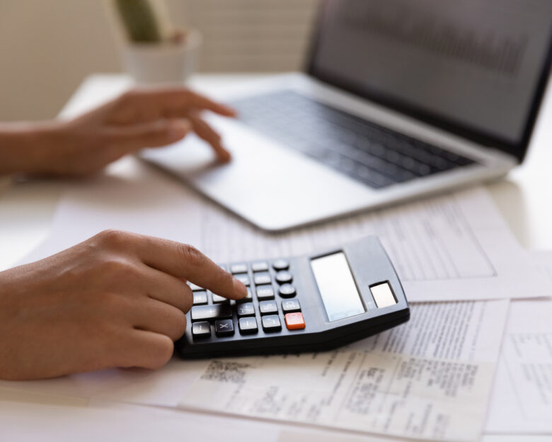 Close up of young female electronic bank client using calculator in paperwork with accounts bills before providing payment online by laptop. Woman hands at work table with papers accounting sum to pay