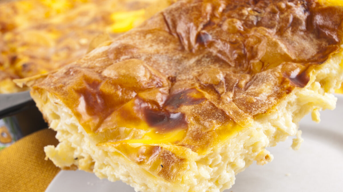 Home made cheese pie in baking tray, one piece on white plate with fork. Yellow tablecloth.