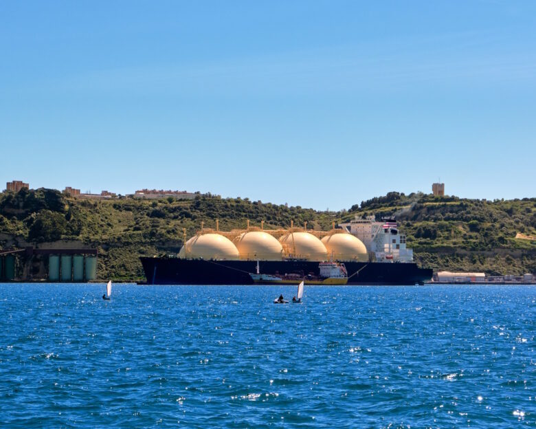 LNG or liquified natural gas tanker moored by industrial pier and green hill on sunny day. Alternative gas supply, commercial freight, energy crisis, regasification, diversification, fuel and energy generation.