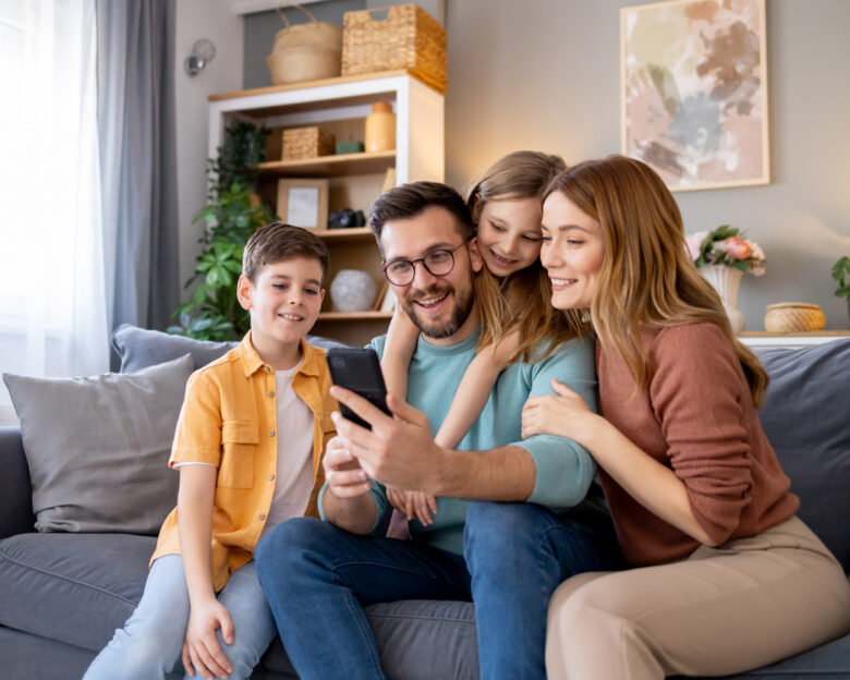 Capturing a cosy, home scene where a happy family with a mother, father, and kids engaging with a mobile phone, enjoying their time together
