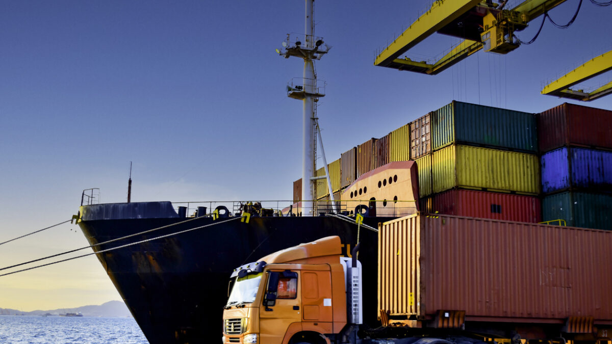 Truck carrying forty-foot container leaving port terminal with ship and quay crane on the background. Seaport operation activities, container shipping, and logistics.