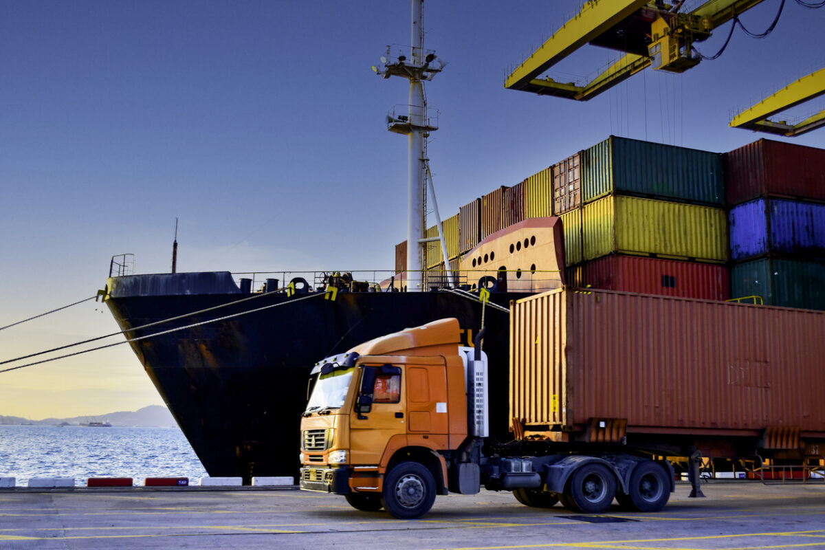 Truck carrying forty-foot container leaving port terminal with ship and quay crane on the background. Seaport operation activities, container shipping, and logistics.