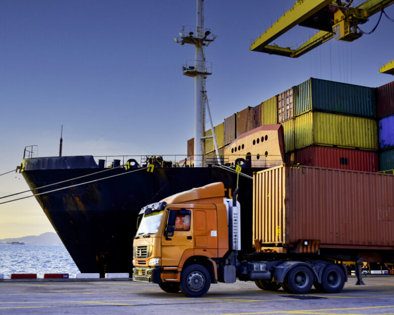 Truck carrying forty-foot container leaving port terminal with ship and quay crane on the background. Seaport operation activities, container shipping, and logistics.
