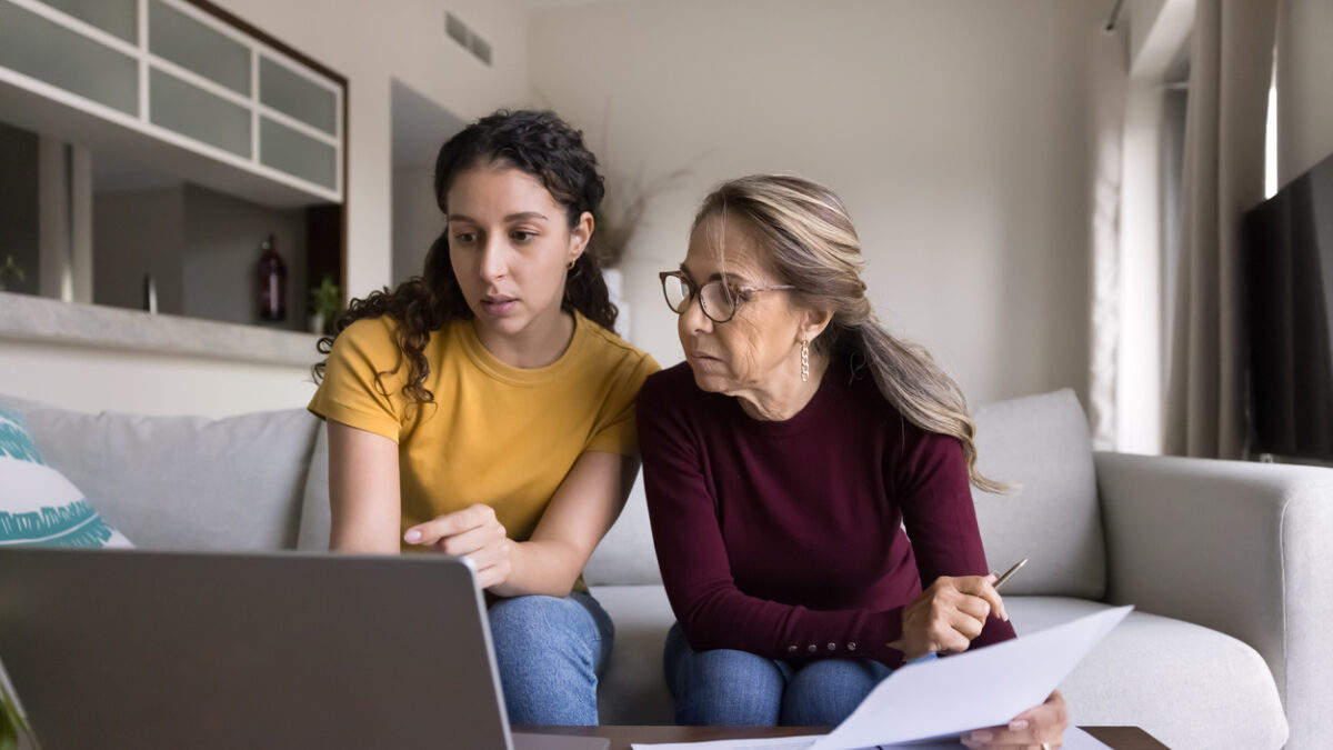 Focused Latin young daughter helping senior mother, showing how to use online financial e-bank application on laptop, paying bills on Internet, checking domestic expenses, budget, fees