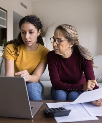 Focused Latin young daughter helping senior mother, showing how to use online financial e-bank application on laptop, paying bills on Internet, checking domestic expenses, budget, fees