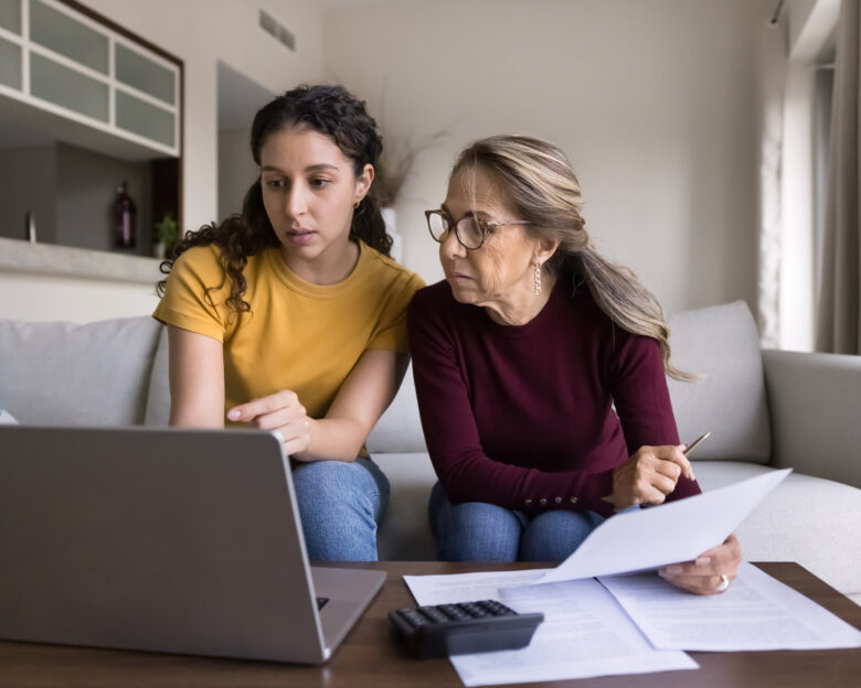Focused Latin young daughter helping senior mother, showing how to use online financial e-bank application on laptop, paying bills on Internet, checking domestic expenses, budget, fees