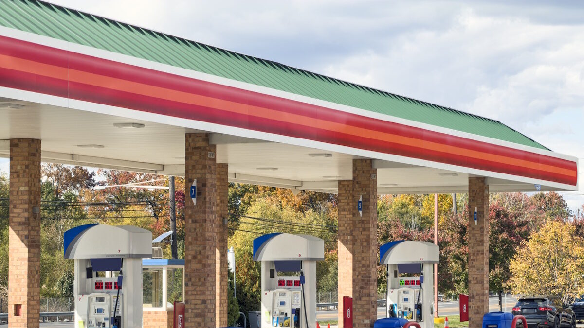 Wide view of a modern gas station with multiple fuel pumps arranged in a row beneath a red, green, and white canopy, set against a clear sky and autumn trees. This full frame transportation and energy image has no copy space and highlights everyday car travel, fuel, and infrastructure, ideal for illustrating concepts of gasoline, service stations, road trips, and automotive industry themes.