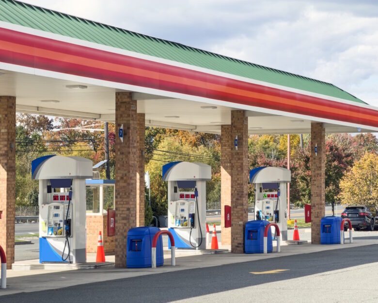 Wide view of a modern gas station with multiple fuel pumps arranged in a row beneath a red, green, and white canopy, set against a clear sky and autumn trees. This full frame transportation and energy image has no copy space and highlights everyday car travel, fuel, and infrastructure, ideal for illustrating concepts of gasoline, service stations, road trips, and automotive industry themes.