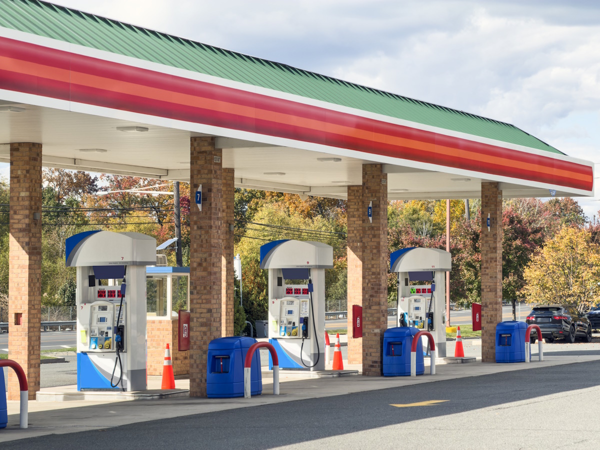 Wide view of a modern gas station with multiple fuel pumps arranged in a row beneath a red, green, and white canopy, set against a clear sky and autumn trees. This full frame transportation and energy image has no copy space and highlights everyday car travel, fuel, and infrastructure, ideal for illustrating concepts of gasoline, service stations, road trips, and automotive industry themes.