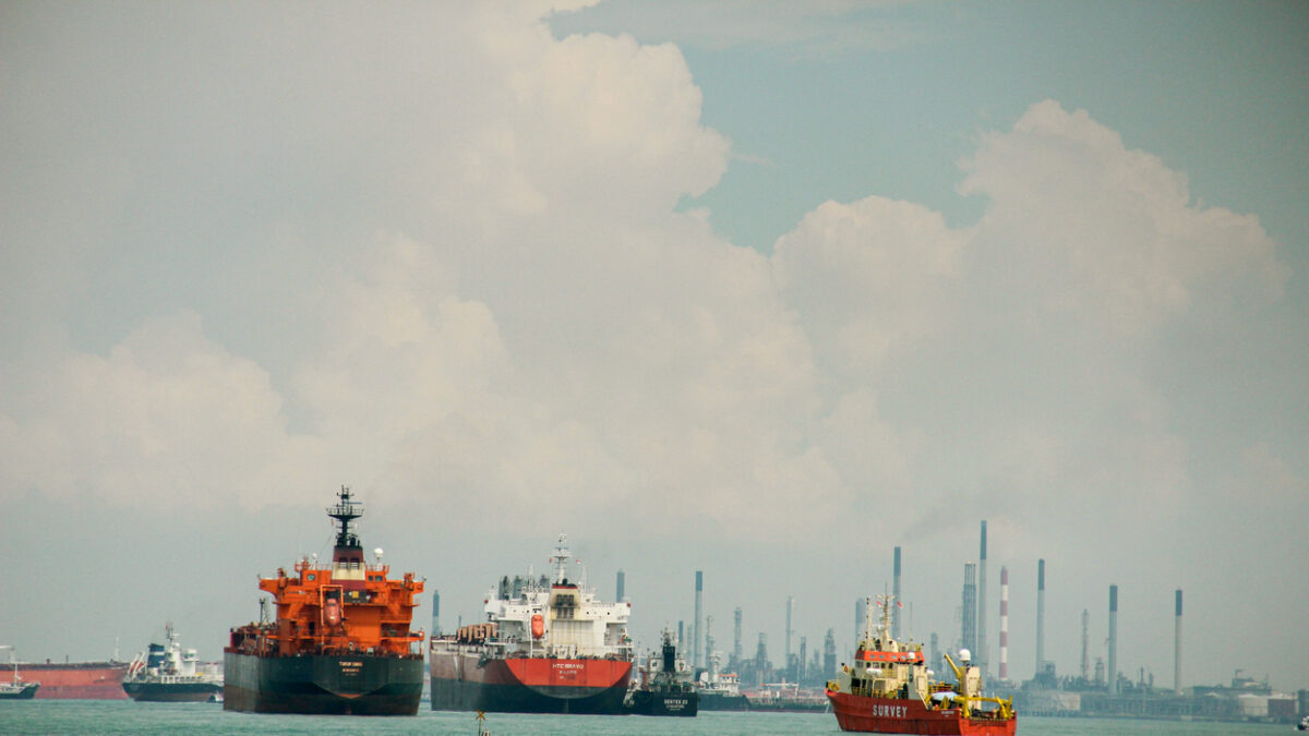 A wide shot of several large commercial vessels, including oil tankers and a survey boat, floating on turquoise water. In the background, an industrial shoreline with numerous smokestacks and refinery infrastructure is visible under a cloudy sky. Concept of global logistics, maritime industry, and international trade.