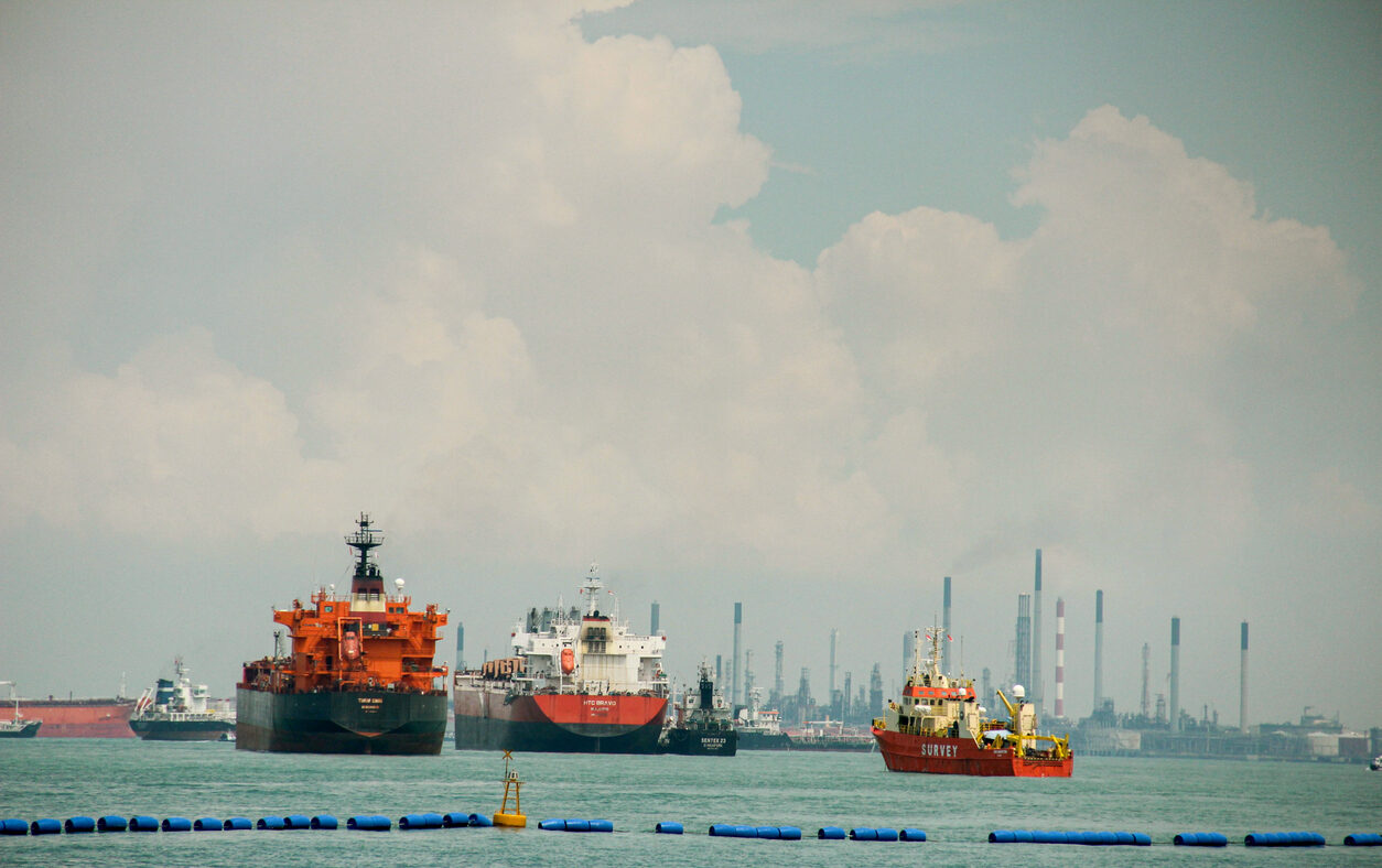 A wide shot of several large commercial vessels, including oil tankers and a survey boat, floating on turquoise water. In the background, an industrial shoreline with numerous smokestacks and refinery infrastructure is visible under a cloudy sky. Concept of global logistics, maritime industry, and international trade.