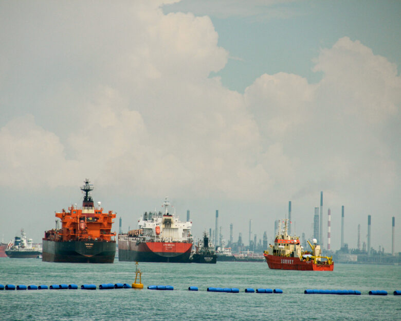 A wide shot of several large commercial vessels, including oil tankers and a survey boat, floating on turquoise water. In the background, an industrial shoreline with numerous smokestacks and refinery infrastructure is visible under a cloudy sky. Concept of global logistics, maritime industry, and international trade.
