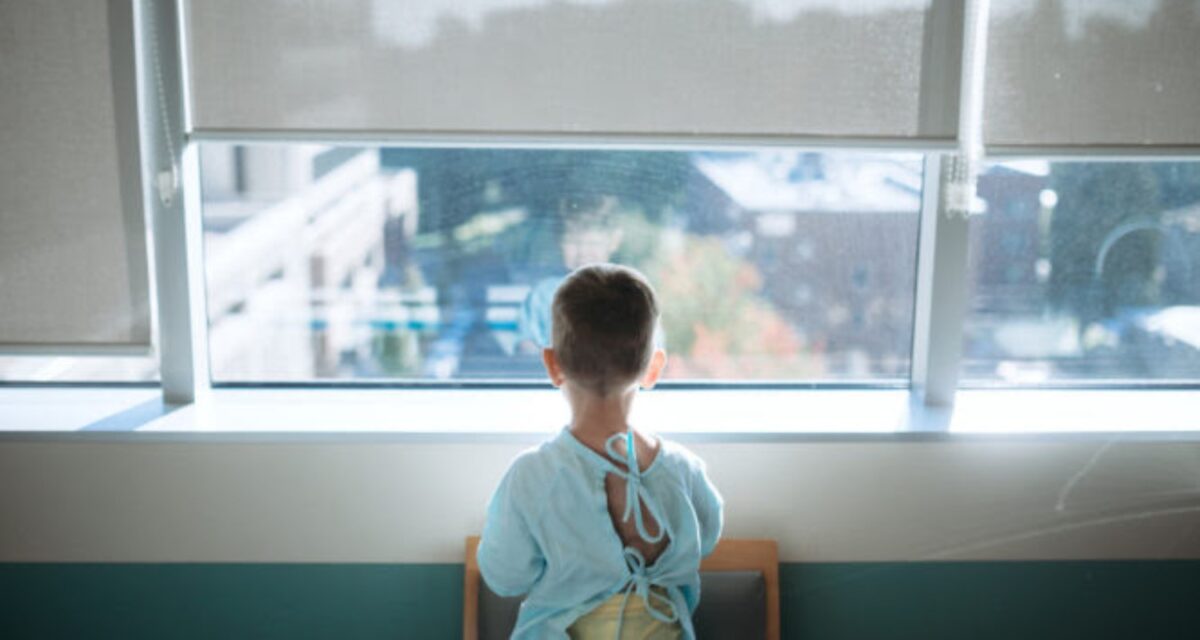 A cute little Caucasian boy waits in the childrens hospital ward to be taken back for surgery. He looks out of the window of his room while waiting for the doctor to arrive.