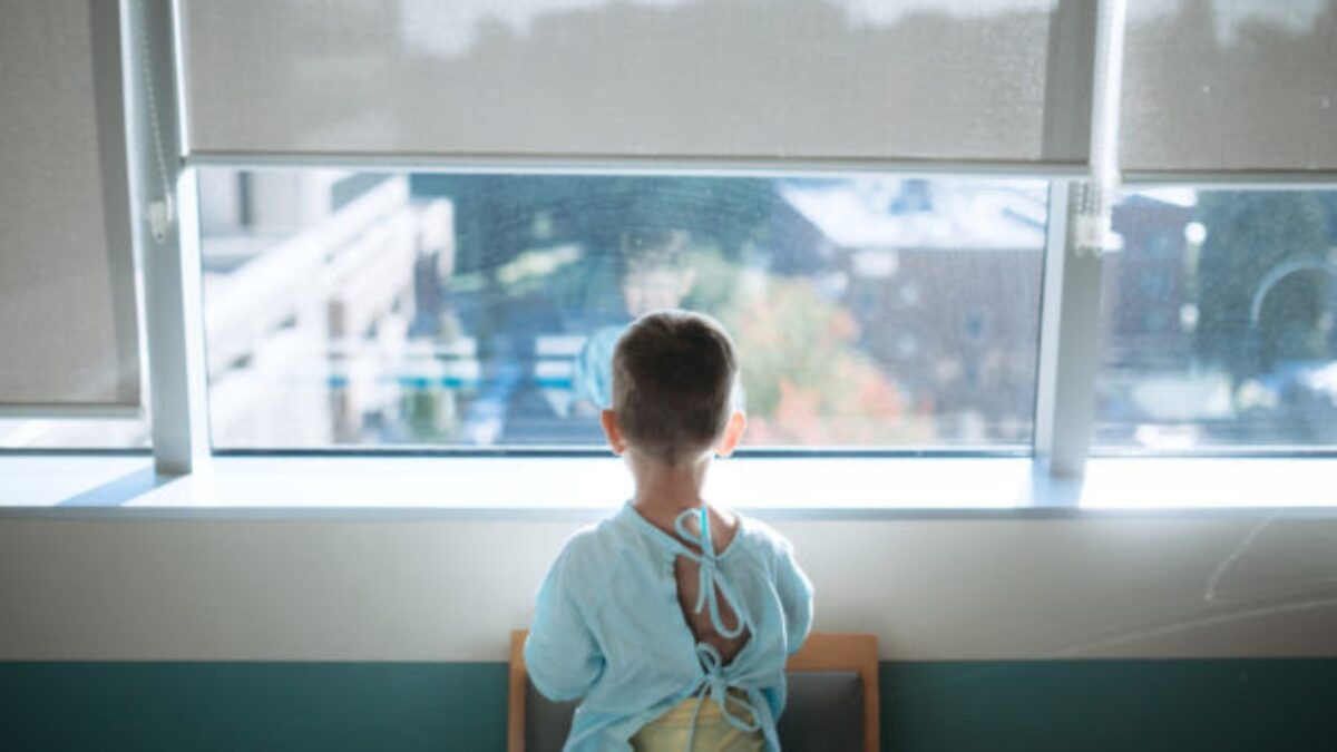 A cute little Caucasian boy waits in the childrens hospital ward to be taken back for surgery. He looks out of the window of his room while waiting for the doctor to arrive.