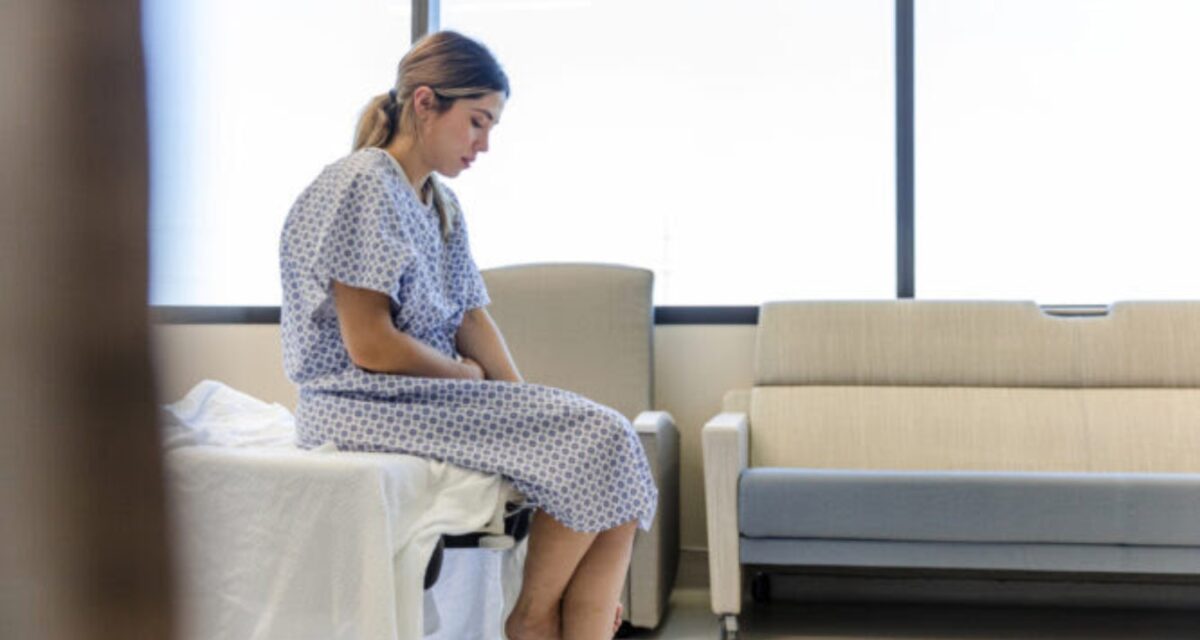 The anxious, sad, young female patient wears her gown as she waits in the hospital room.
