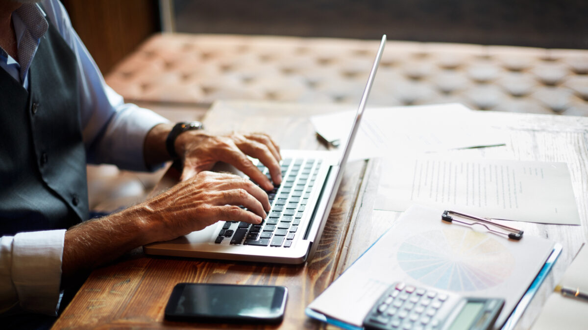 Elderly unrecognizable entrepreneur using a laptop. Mobile phone, a calculator, a pie chart and some papers and notes are on the table.