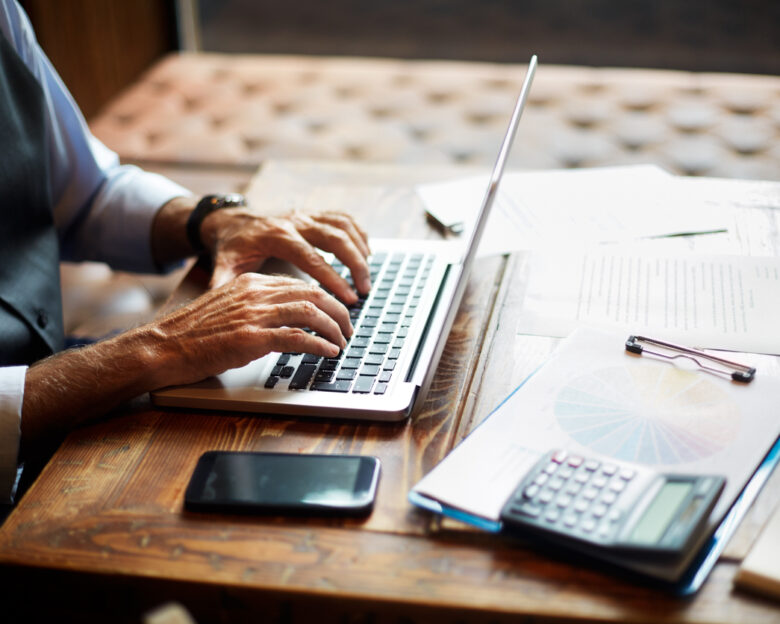 Elderly unrecognizable entrepreneur using a laptop. Mobile phone, a calculator, a pie chart and some papers and notes are on the table.