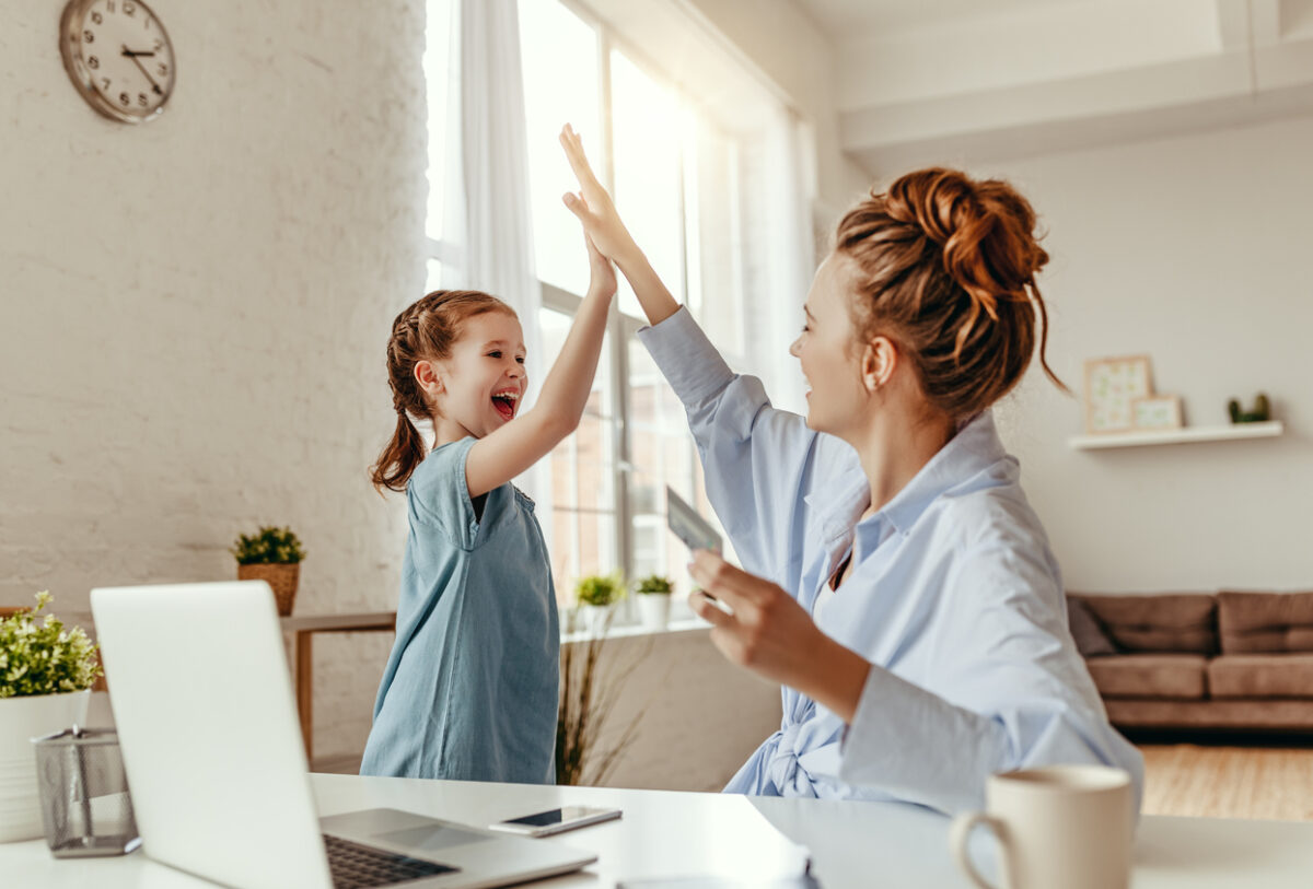 Low angle of excited small daughter giving high five to mother and screaming while celebrating successful online shopping using laptop at table in light living room