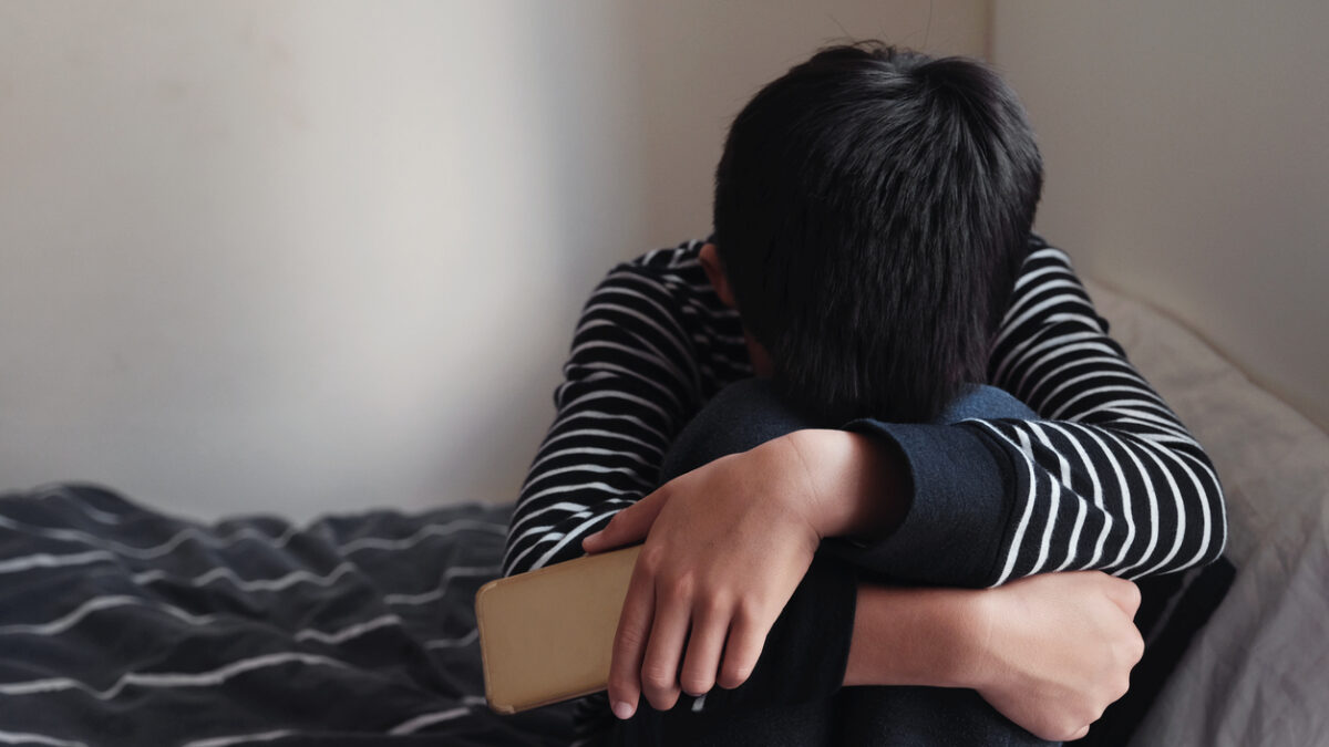 oung Asian preteen teenager boy hugging his knee in his bedroom with smartphone, Cyber bullying in kid, depressed child mental health
