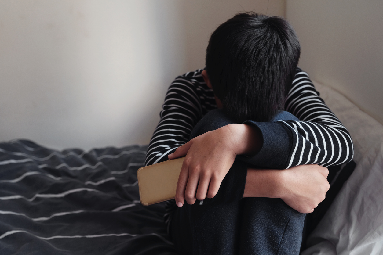 oung Asian preteen teenager boy hugging his knee in his bedroom with smartphone, Cyber bullying in kid, depressed child mental health