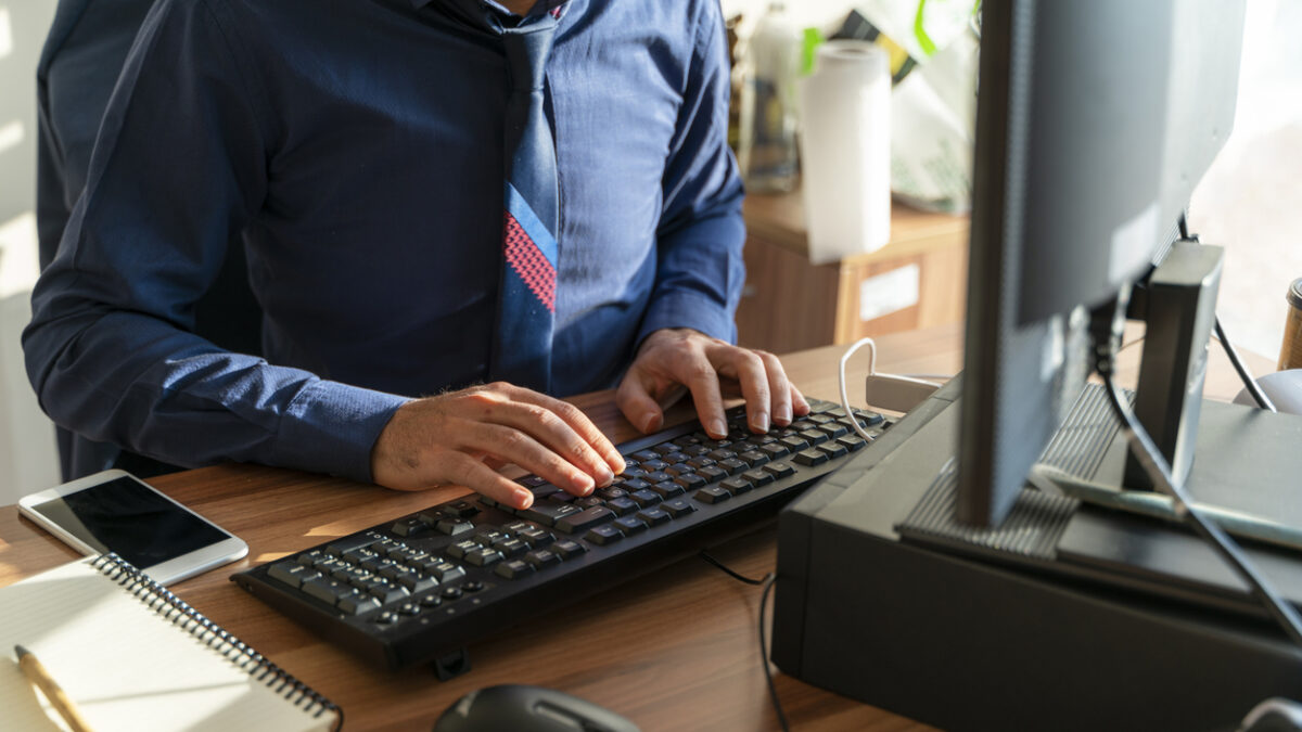 A background image of an office worker at a desk. The image, taken with a full-frame camera, shows a desktop computer, keyboard, notebook, cell phone, computer mouse, and a hand typing on the keyboard