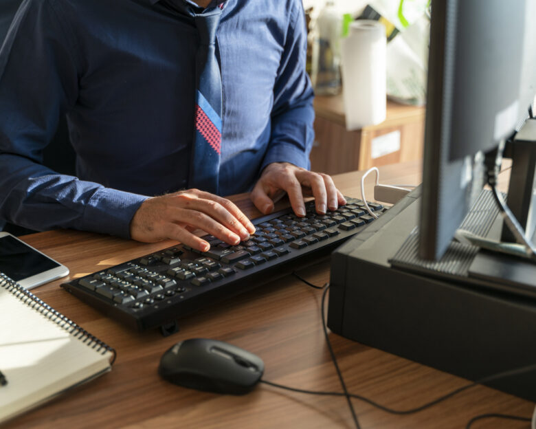 A background image of an office worker at a desk. The image, taken with a full-frame camera, shows a desktop computer, keyboard, notebook, cell phone, computer mouse, and a hand typing on the keyboard