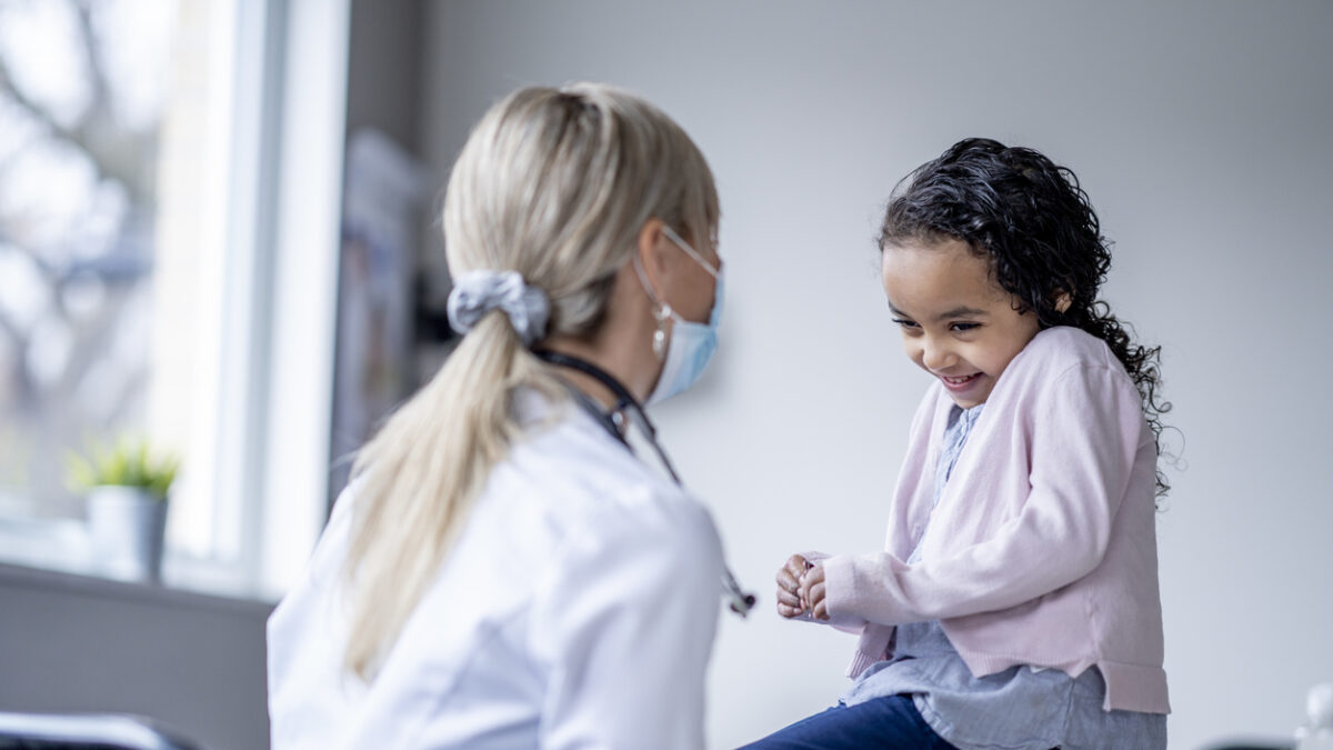 A sweet little girl sits up on an exam table nervously giggling during an appointment. She is dressed casually in a pale pink sweater as she gazes at the doctor. The female doctor is wearing a white lab coat, as a stethoscope around her neck and a medical face mask on as she tries to put the little girl at ease and make the appointment fun.