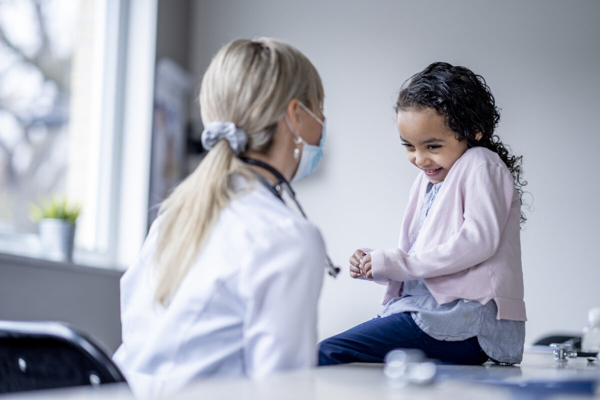 A sweet little girl sits up on an exam table nervously giggling during an appointment. She is dressed casually in a pale pink sweater as she gazes at the doctor. The female doctor is wearing a white lab coat, as a stethoscope around her neck and a medical face mask on as she tries to put the little girl at ease and make the appointment fun.