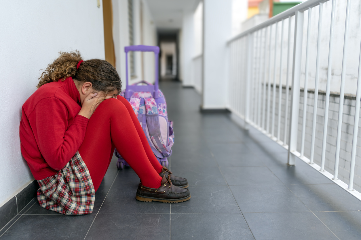 Girl in school uniform with backpack alone crying sitting on the floor in the school hallway. Bullying, rejection and isolation. Adhd and autism.