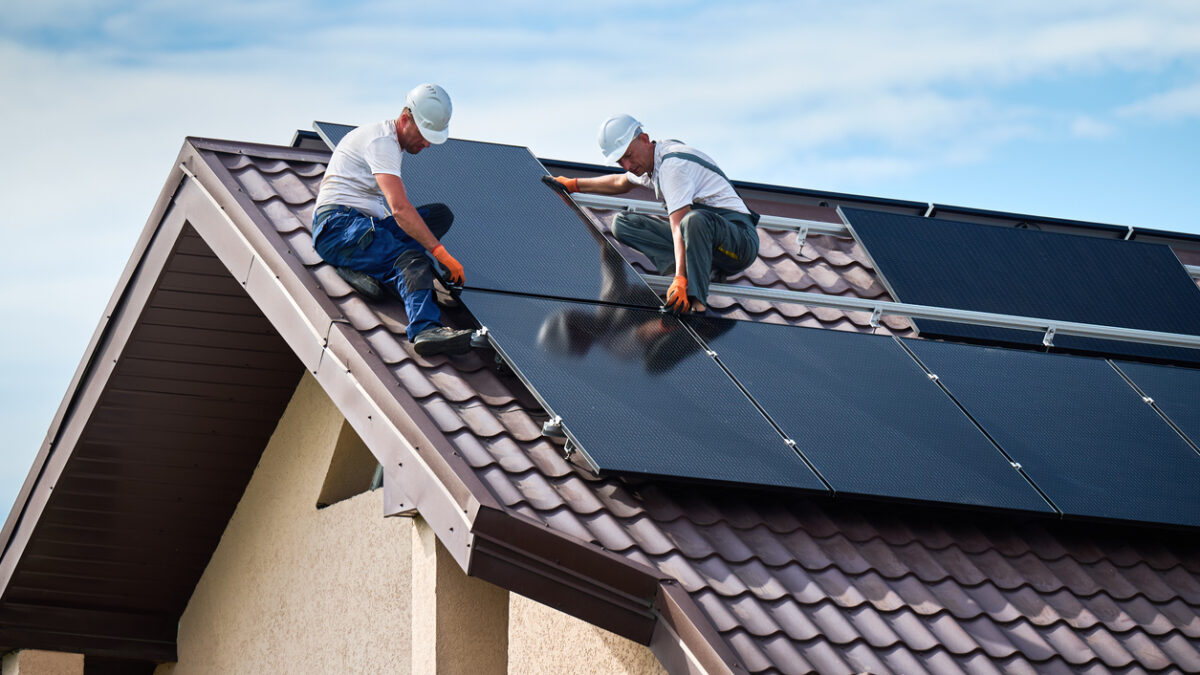 Workers building solar panel system on rooftop of house. Two men installers in helmets installing photovoltaic solar module outdoors. Alternative, green and renewable energy generation concept.