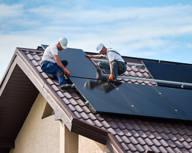 Workers building solar panel system on rooftop of house. Two men installers in helmets installing photovoltaic solar module outdoors. Alternative, green and renewable energy generation concept.