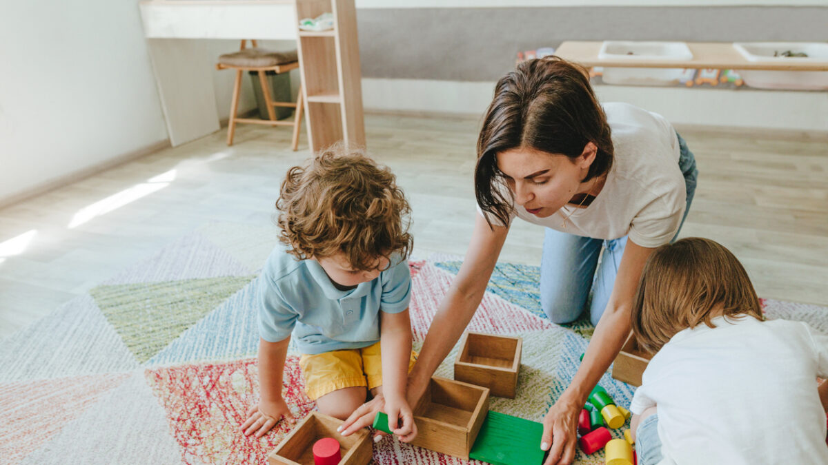 Young woman teacher playing in a kindergarten with children on the floor with wooden multi-colored toys. Selective focus.