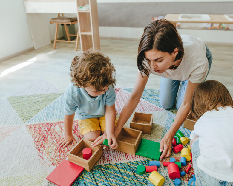 Young woman teacher playing in a kindergarten with children on the floor with wooden multi-colored toys. Selective focus.