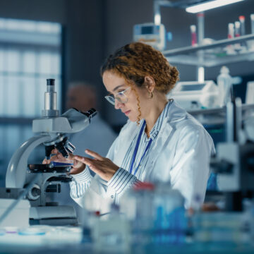 Hisplanic Female Bioengineer Looking at DNA Samples Under a Microscope in a Modern Applied Science Laboratory. Portrait of a Young Lab Engineer in White Coat Inventing New Medical Drugs