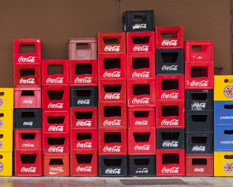 Group of stacks of multi colored plastic crates for beverage bottles, seen on a town street sidewalk outside a bar. Several beer, soda and soft drink brands on them. Concrete wall in the background.
