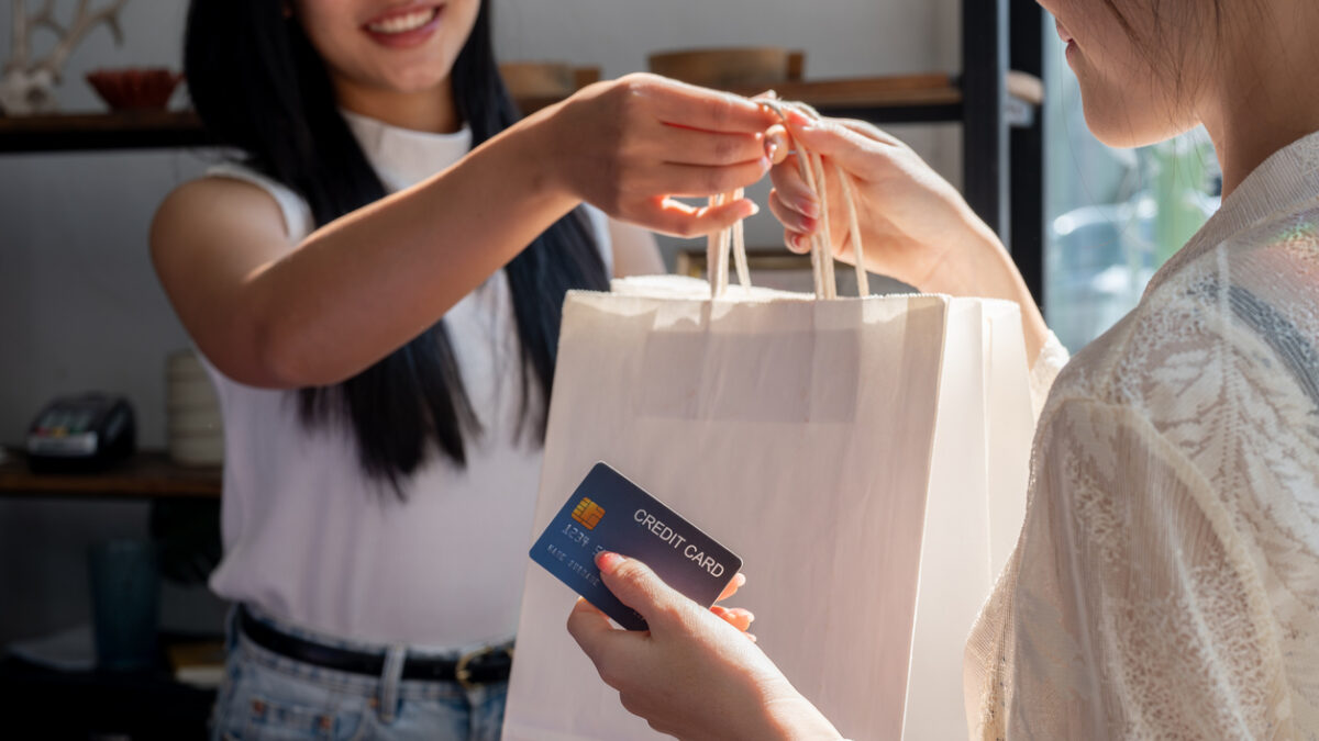A close-up of a friendly Asian female shop cashier warmly smiling as she hands shopping bags to a customer after checkout, working in a retail store.