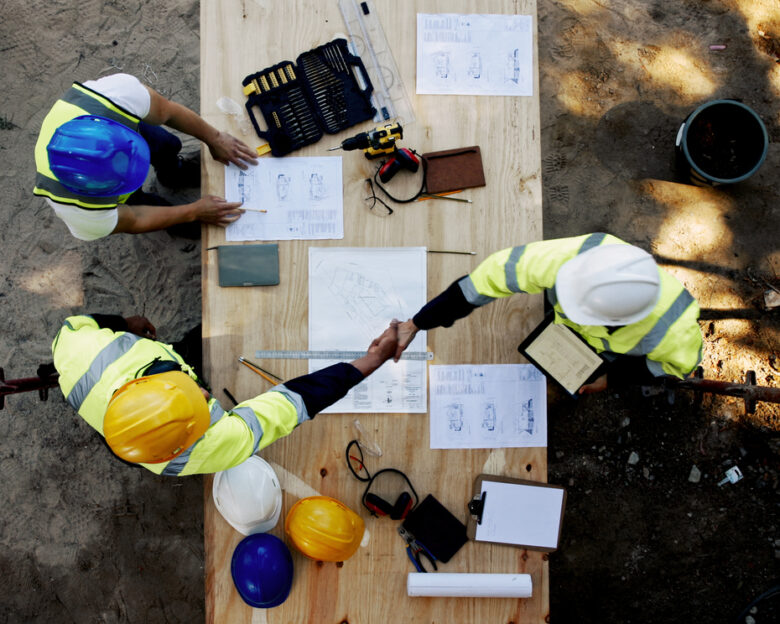 Blueprint, architecture and men at table shaking hands for collaboration, agreement and inspection from above. Construction, building team and contractor on site with floor plan, handshake and deal