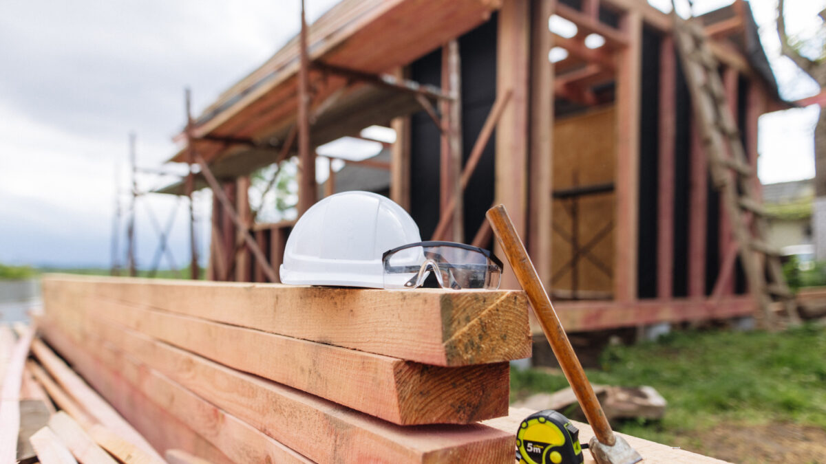 The protective helmet, glasses and hammer lie on the stacked lumber at the frame house construction site
